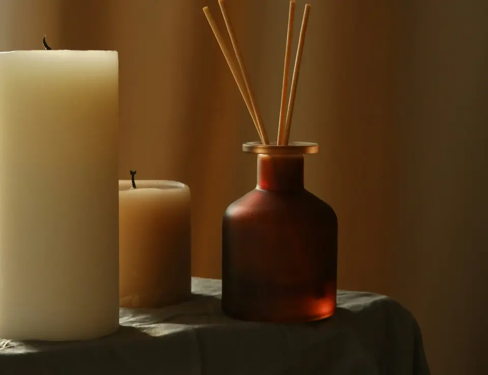 reed diffuser in amber coloured glass bottle placed next to two tower white candles