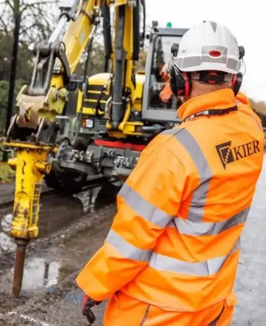 a man in an orange jacket and white helmet standing beside a yellow machine