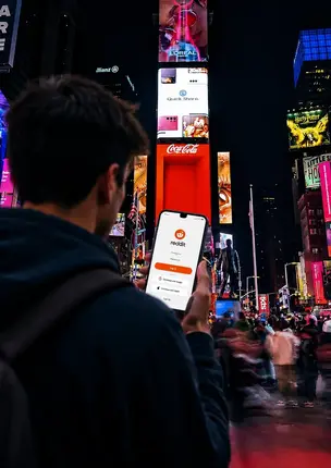 Man holding phone in Times Square, the phone screen shows the Reddit login page 